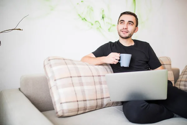 El hombre pensativo con el portátil y la taza de café en el sofá en casa — Foto de Stock