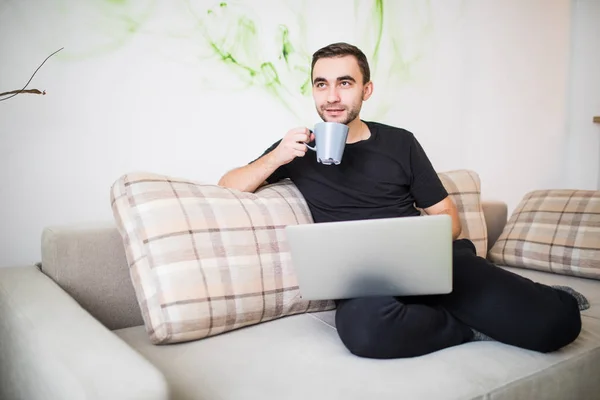 Feliz joven sentado en el sofá con portátil y auriculares y sosteniendo una taza de café en su mano . — Foto de Stock