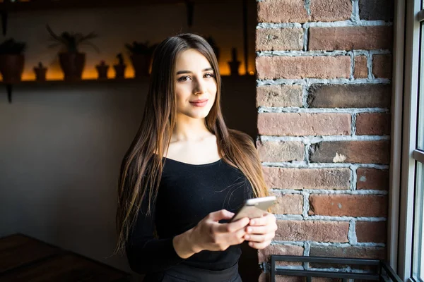 Portrait of a cheerful woman holding smartphone texting and looking at window in cafe shop — Stock Photo, Image