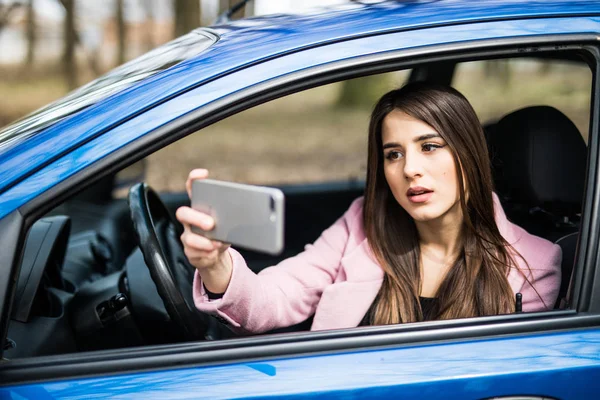Улыбающаяся молодая женщина делает селфи с камерой смартфона на улице в синей машине — стоковое фото