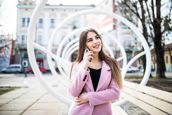 Mulher alegre falando ao telefone na rua perto do banco moderno usando uma jaqueta rosa — Fotografia de Stock