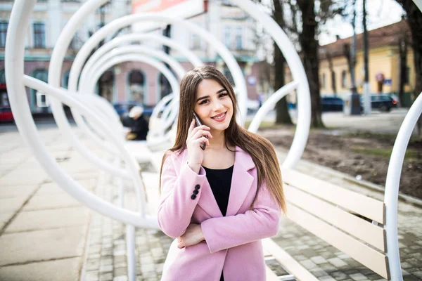 Primer plano retrato de chica morena disfrutada hablando por teléfono en la calle cerca del banco moderno. Ella lleva chaqueta rosa, sonriendo a un lado —  Fotos de Stock