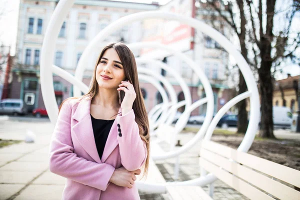 Primer plano retrato de chica morena disfrutada hablando por teléfono en la calle cerca del banco moderno. Ella lleva chaqueta rosa, sonriendo a un lado —  Fotos de Stock