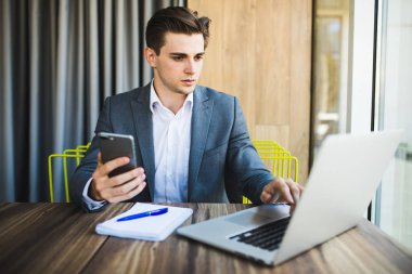 Genç mutlu iş adamı akıllı telefonunu okurken gülümsüyor. Ofiste akıllı telefonla mesaj okuyan gülümseyen iş adamının portresi. Ofisteki masasında çalışan bir adam..