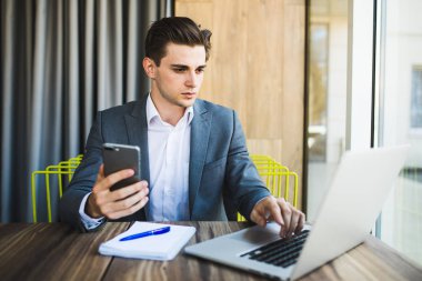 Genç mutlu iş adamı akıllı telefonunu okurken gülümsüyor. Ofiste akıllı telefonla mesaj okuyan gülümseyen iş adamının portresi. Ofisteki masasında çalışan bir adam..