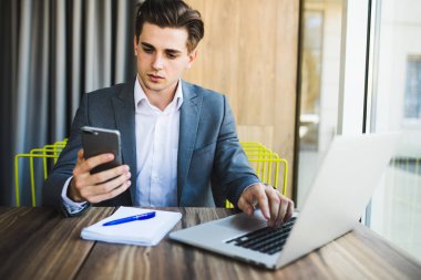 Kendine güvenen genç adam akıllı casual giyim akıllı telefon tutarak ve Office çalışma evinde otururken bakmak