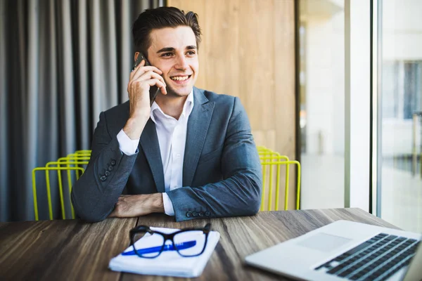 Retrato de joven guapo en gafas sentado en el escritorio de la oficina con ordenador portátil y hablando por teléfono móvil . — Foto de Stock