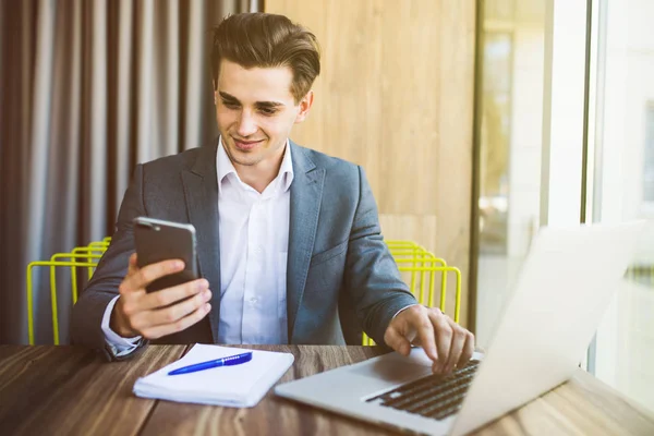 Joven confiado en ropa casual inteligente sosteniendo el teléfono inteligente y mirándolo mientras está sentado en su lugar de trabajo en la oficina — Foto de Stock