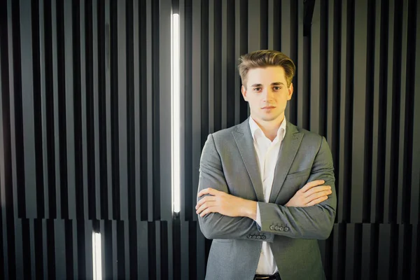 Retrato de un joven guapo en un traje de negocios con las manos cruzadas contra la pared oscura de madera en la oficina moderna — Foto de Stock