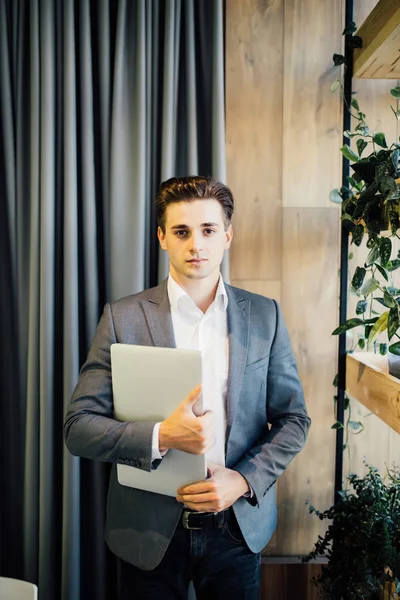 Joven hombre guapo de confianza en camisa y corbata sosteniendo el ordenador portátil y sonriendo mientras está de pie en la oficina moderna — Foto de Stock