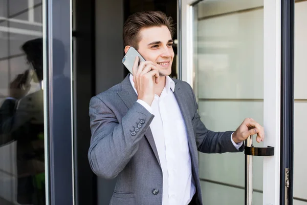 Młody biznesmen mówić na jego telefon w pobliżu drzwi szklane w budynku biurowym — Zdjęcie stockowe