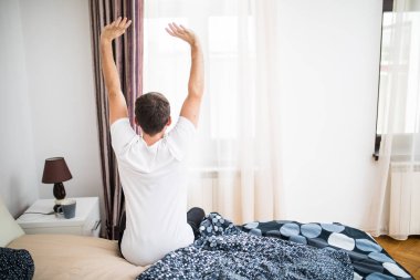 Yatakta uyanma ve kollarını evde, arka görünümünden germe genç adam