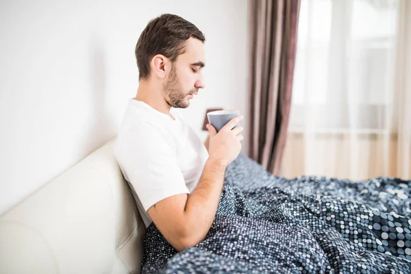Красивый молодой человек пьет кофе в постели дома — стоковое фото