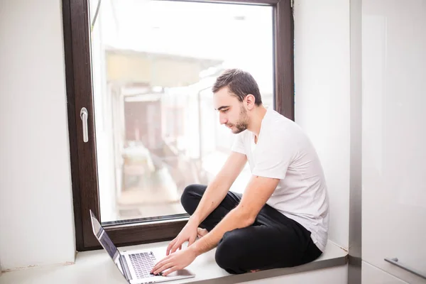 Feliz hombre sonriente sentado en el alféizar de la ventana y escribiendo en el ordenador portátil en casa — Foto de Stock