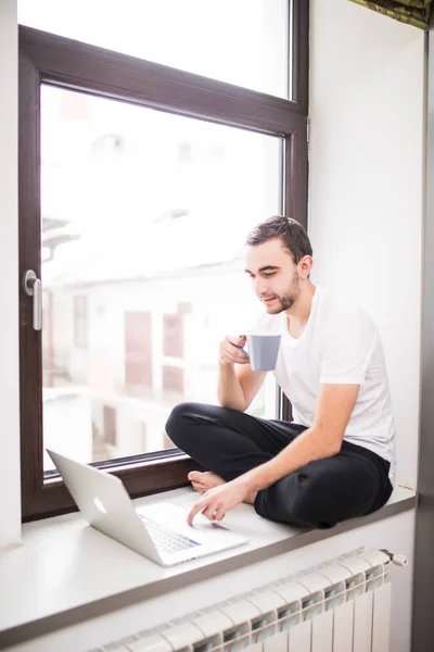 Hombre joven sentado en el alféizar de la ventana con el ordenador portátil, taza de café en casa — Foto de Stock