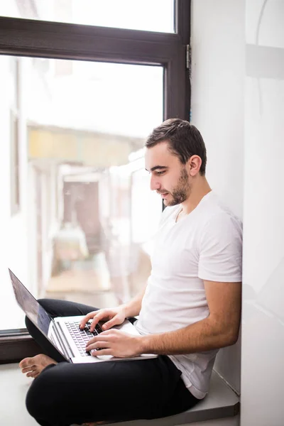 El valiente, guapo, un estudiante trabajando en un alféizar de ventana con un portátil. trabajador que se comunica en Internet . — Foto de Stock