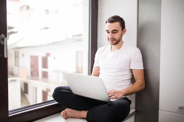 El valiente, guapo, un estudiante trabajando en un alféizar de ventana con un portátil. trabajador que se comunica en Internet . — Foto de Stock