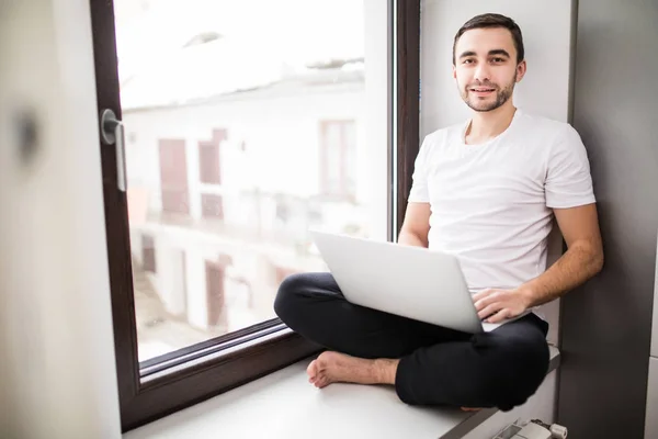 Joven atractivo usando un ordenador portátil, mirando a la cámara y sonriendo mientras estudia en casa — Foto de Stock