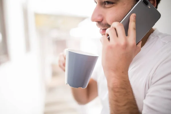 Mladý muž na sobě neformální rozhovor na mobilním telefonu ráno v okně. — Stock fotografie