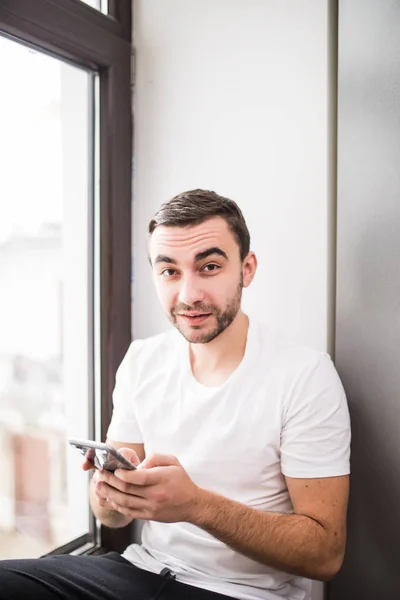 Bir telefon tutarak ve evde iletileri okuma ev ortamında bir pencere üzerinde oturan beyaz adam — Stok fotoğraf