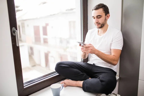 Белый человек сидит на подоконнике в домашней обстановке, держит телефон и читает сообщения дома. — стоковое фото