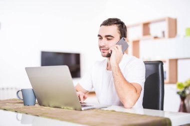 Yakışıklı işadamı cep telefonda konuşurken ve mutfakta bir dizüstü bilgisayar kullanırken gülümseyen