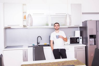 Bir fincan kahve uzağa düşünmeden bakarak tutan mutfakta ayakta adam portresi. Evde mutfakta kahve rahat genç adam