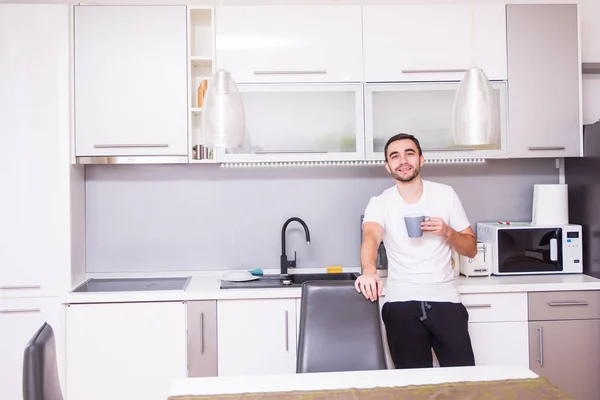 Bir fincan kahve uzağa düşünmeden bakarak tutan mutfakta ayakta adam portresi. Evde mutfakta kahve rahat genç adam — Stok fotoğraf