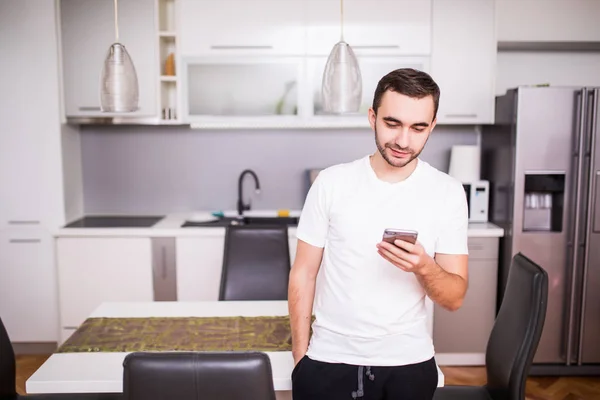 Hombre joven guapo en camisa a cuadros de pie y el uso de teléfono inteligente en la cocina — Foto de Stock