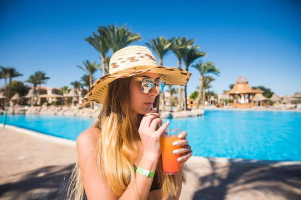 Приваблива усміхнена брюнетка влітку з коктейлем і солом'яним капелюхом біля басейну — стокове фото