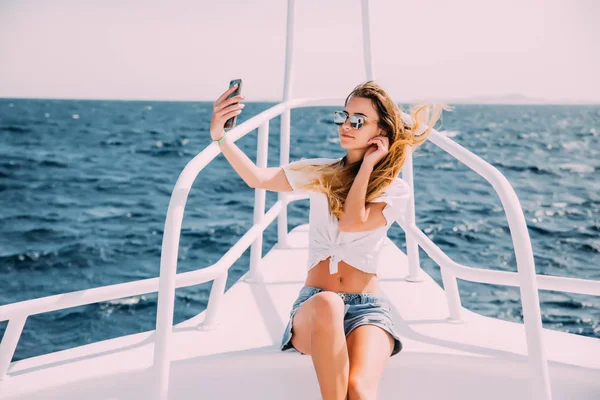 Fiatal lány ül a yacht, és figyelembe selfie a tenger a háttérben — Stock Fotó