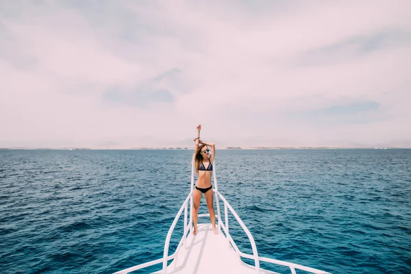 Красивая девушка в черном купальнике, стоящая на носу яхты и расправляющая руки в солнечный летний день на морском фоне — стоковое фото