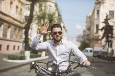 Kask içinde neşeli genç sakallı adam scooter üzerinde oturuyor ve caddenin üzerinde Merhaba deyin. tonda.