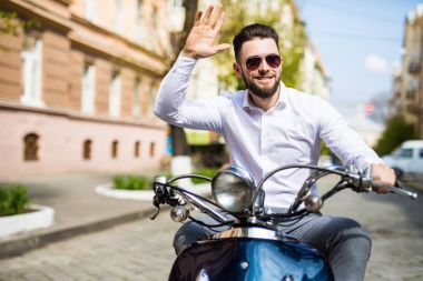 Cheerful young bearded man in helmet is sitting on scooter and say hello on city street clipart