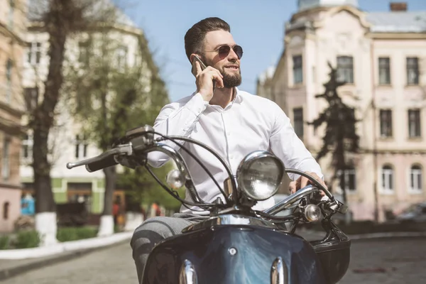 Веселый молодой человек, сидящий на скутере и разговаривающий по мобильному телефону. тонированные — стоковое фото