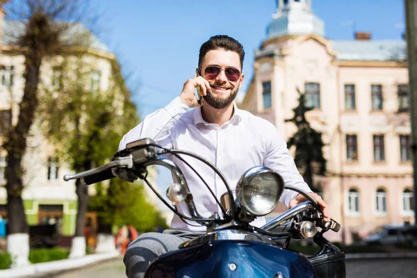 Příjemný mladý muž sedí na skútru a mluví na mobilním telefonu — Stock fotografie