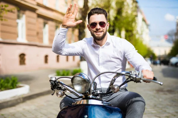 Jovem barbudo alegre homem no capacete está sentado em scooter e dizer olá na rua da cidade — Fotografia de Stock