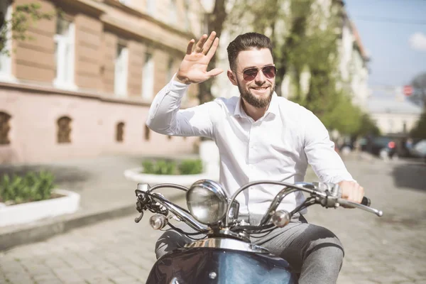 Jovem barbudo alegre homem no capacete está sentado na scooter e dizer olá na rua da cidade. tonificado . — Fotografia de Stock