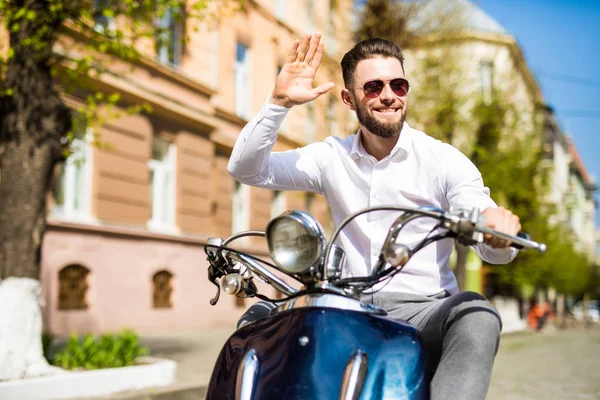 Jovem barbudo alegre homem no capacete está sentado em scooter e dizer olá na rua da cidade — Fotografia de Stock