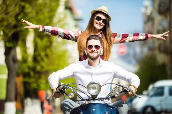 Beautiful young couple riding scooter together while happy woman raising arms and smiling