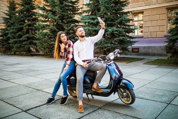 Красивая молодая любящая пара, сидящая вместе на скутере и делающая селфи со своего смартфона — стоковое фото