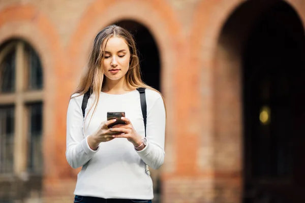 Hermosa estudiante feliz sosteniendo portátiles mensajes de texto en el teléfono en el campus en la universidad — Foto de Stock