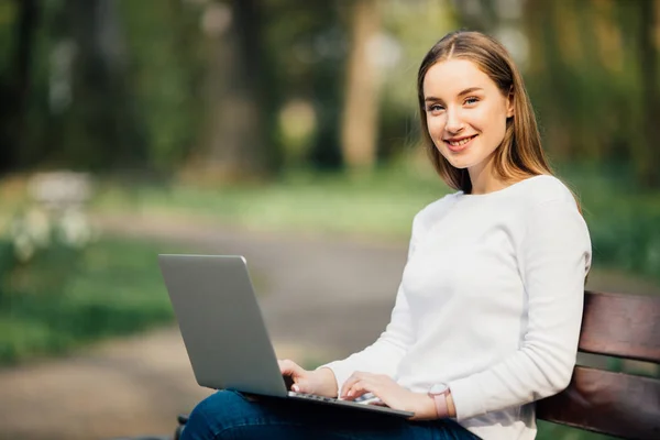 Молодая элегантная женщина сидит на скамейке в парке и работает на ноутбуке — стоковое фото