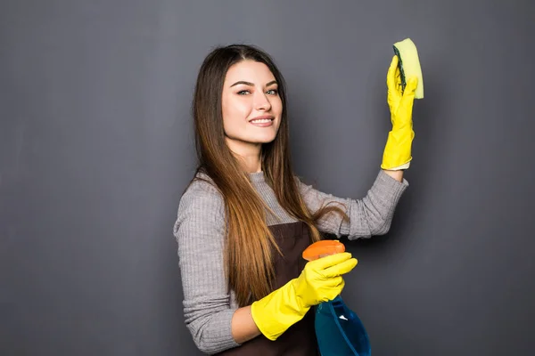 Молода жінка використовує спрей під час прибирання будинку позує на камеру на сірому фоні — стокове фото
