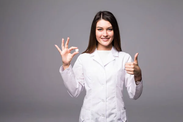 Wesoły atrakcyjna młoda kobieta lekarz trzymając tabletkę i pokazuje kciuk w górę na szarym tle. — Zdjęcie stockowe