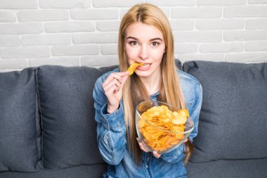 Portrait of smiling girl eating chips at home on sofa clipart