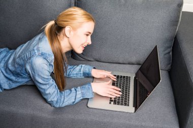 Casual young woman lying on couch and using laptop at home clipart