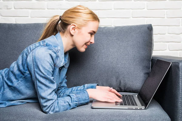 カジュアルな若い女性のソファに横たわっていると自宅でラップトップを使用して — ストック写真