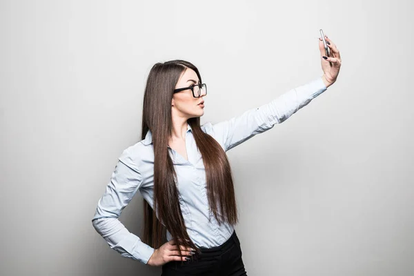 Приваблива молода жінка в повсякденному одязі робить селфі на смартфоні і посміхається, стоячи на сірому фоні — стокове фото