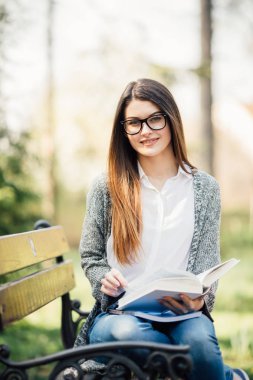 kitap gülümseyen ve kameraya bakarak onun elinde tutan banktaoturuyor, güzel ve mutlu genç öğrenci kız.